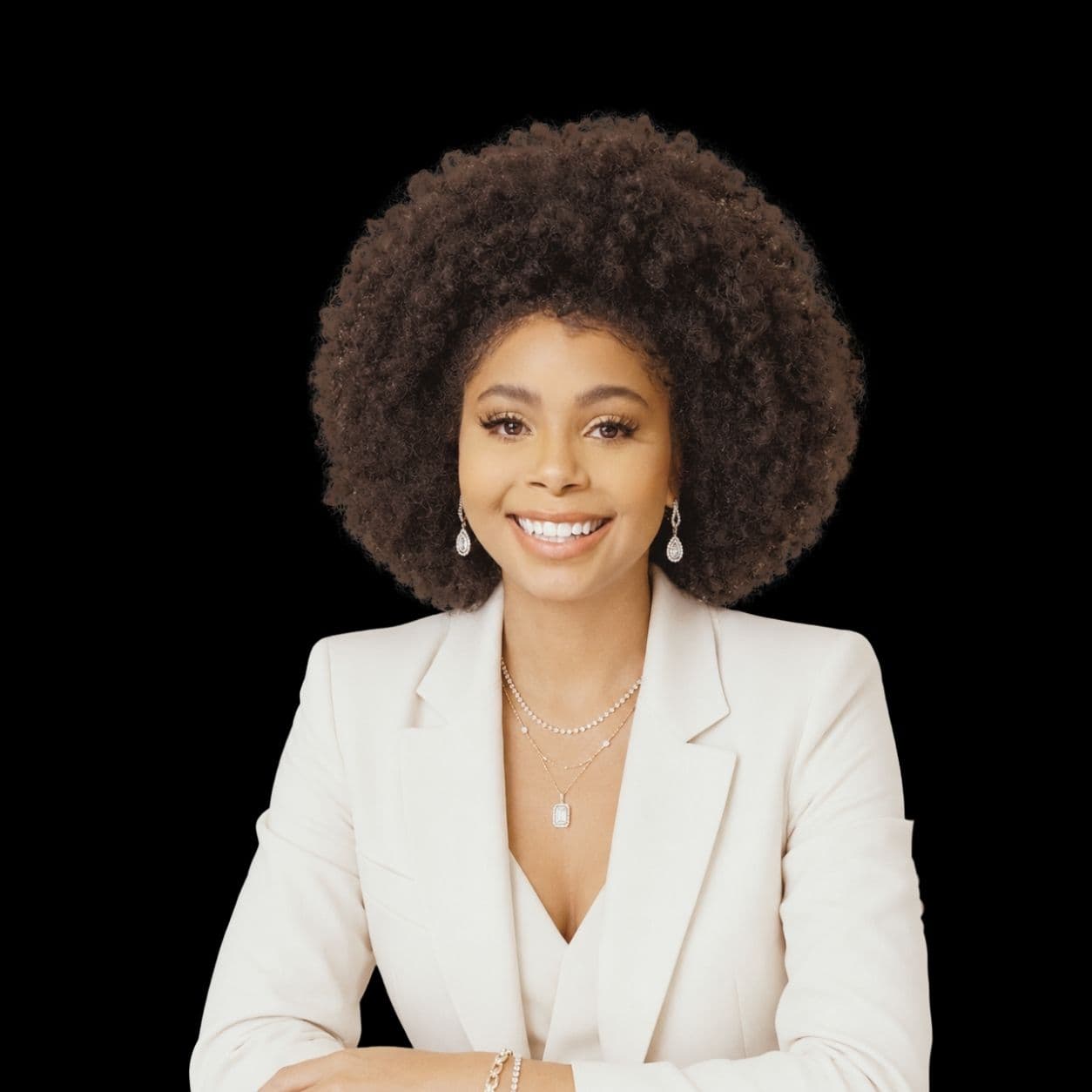 Smiling woman with a large afro hairstyle wearing a white blazer and elegant jewelry.